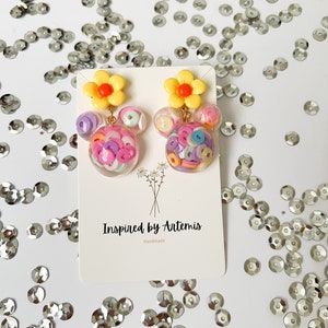 May include: A pair of handmade earrings featuring a yellow flower charm with a red center and a clear resin sphere filled with colorful confetti. The earrings are displayed on a white card with the text "Inspired by Artemis Handmade".