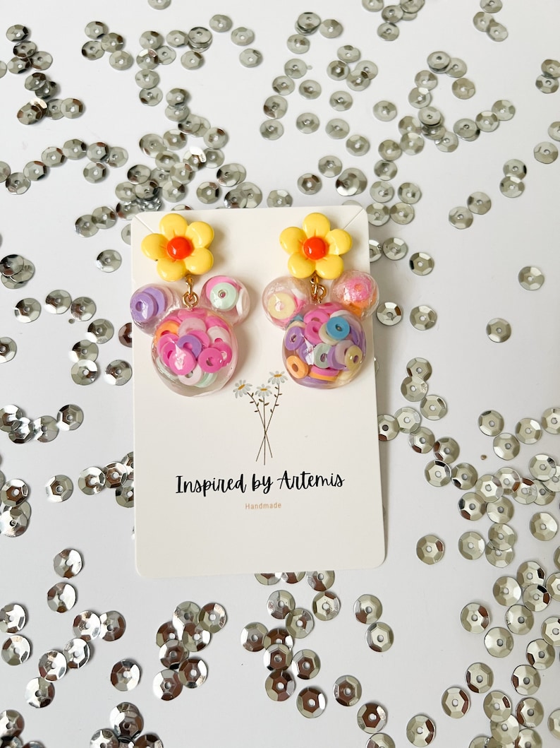 May include: A pair of handmade earrings featuring a yellow flower charm with a red center and a clear resin sphere filled with colorful confetti. The earrings are displayed on a white card with the text "Inspired by Artemis Handmade".