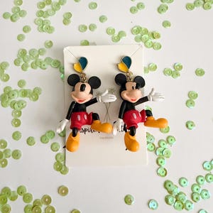 May include: A pair of Mickey Mouse earrings. The earrings feature a black head with large ears, a red shorts, yellow shoes, and white gloves. The earrings are on a white card with the word "inspiring" and "handmade" printed on it. Green sequins are scattered around the earrings.