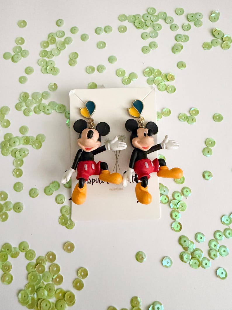 May include: A pair of Mickey Mouse earrings. The earrings feature a black head with large ears, a red shorts, yellow shoes, and white gloves. The earrings are on a white card with the word "inspiring" and "handmade" printed on it. Green sequins are scattered around the earrings.