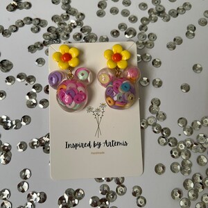 May include: A pair of colorful earrings with a yellow flower charm and a clear sphere filled with colorful confetti. The earrings are handmade and are labeled "Inspired by Artemis".