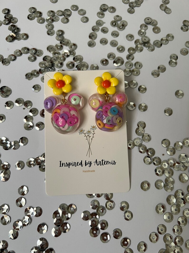 May include: A pair of colorful earrings with a yellow flower charm and a clear sphere filled with colorful confetti. The earrings are handmade and are labeled "Inspired by Artemis".