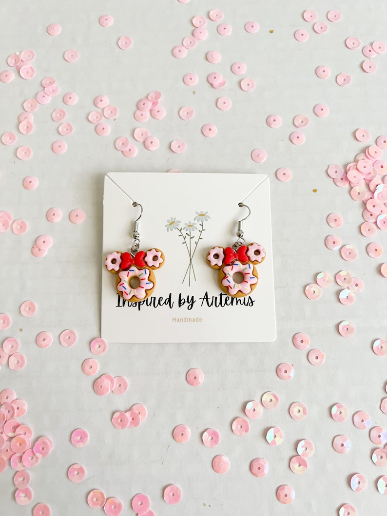 May include: A pair of donut-shaped earrings with pink frosting and sprinkles, red bows, and silver hooks. The earrings are displayed on a white card with the text "Inspired by Artemis" and "Handmade".