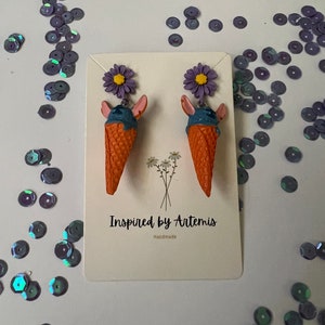 May include: A pair of handmade ice cream cone earrings with purple flowers and blue and orange details. The earrings are displayed on a white card with the text "Inspired by Artemis" and "Handmade".