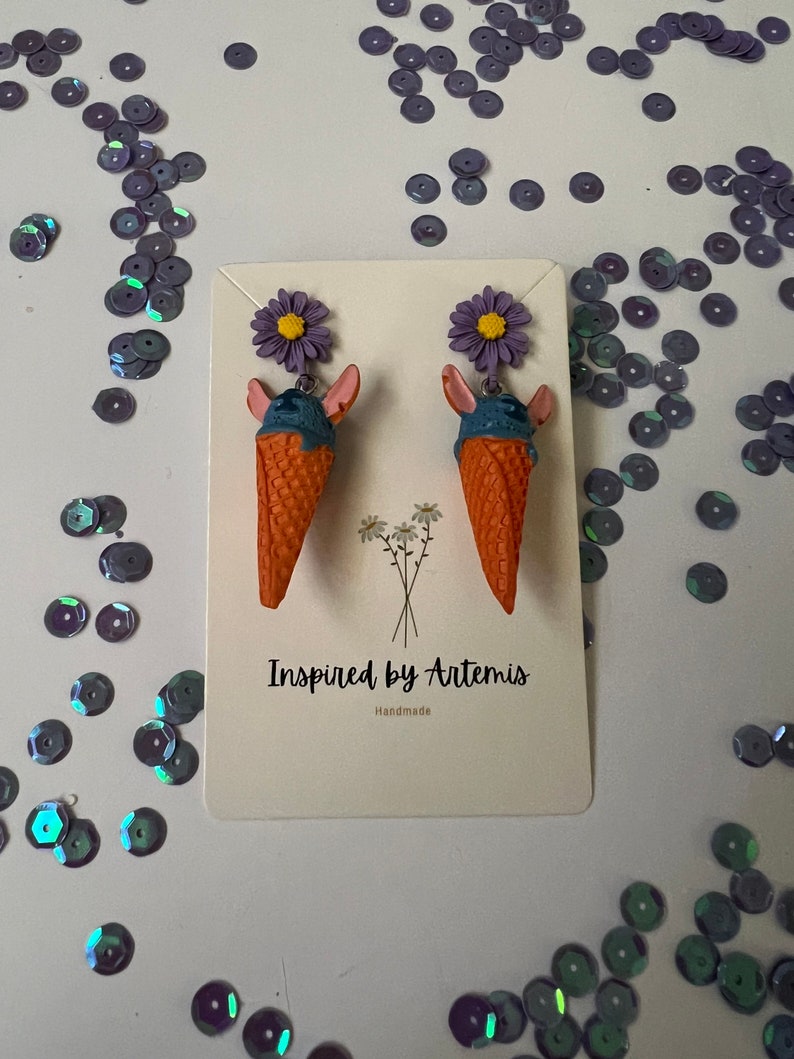 May include: A pair of handmade ice cream cone earrings with purple flowers and blue and orange details. The earrings are displayed on a white card with the text "Inspired by Artemis" and "Handmade".