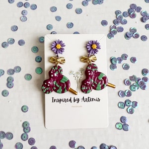 May include: Purple and green swirl lollipop earrings with a gold bow and a purple flower charm. The earrings are handmade and the card says "Inspired by Artemis".