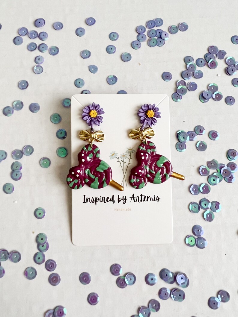 May include: Purple and green swirl lollipop earrings with a gold bow and a purple flower charm. The earrings are handmade and the card says "Inspired by Artemis".
