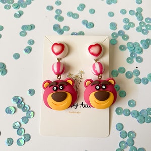 May include: A pair of dangle earrings featuring a pink heart, a pink and white striped ball, and a pink bear face with brown eyes and a yellow smile. The earrings are handmade and are displayed on a white card with the text "by Ari" and "Handmade".