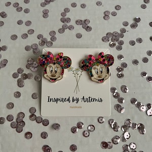 May include: A pair of gold-tone earrings featuring a cartoon character with a pink bow and colorful rhinestones. The earrings are on a white card with the text "Inspired by Artemis" and "Handmade".