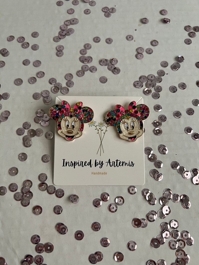 May include: A pair of gold-tone earrings featuring a cartoon character with a pink bow and colorful rhinestones. The earrings are on a white card with the text "Inspired by Artemis" and "Handmade".