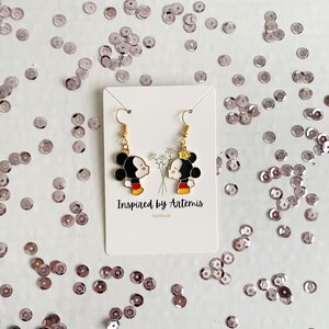 May include: A pair of gold-tone earrings featuring a cartoon couple kissing. The earrings are on a white card with the text "Inspired by Artemis" and "Handmade".