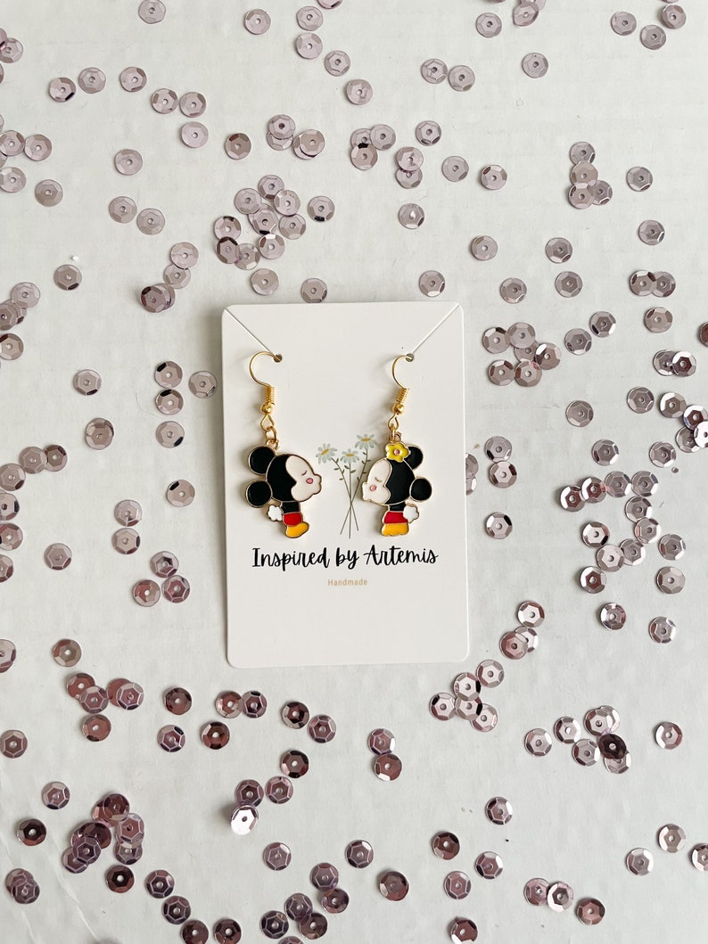 May include: A pair of gold-tone earrings featuring a cartoon couple kissing. The earrings are on a white card with the text "Inspired by Artemis" and "Handmade".