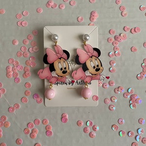May include: Pink Minnie Mouse earrings with a pink bow and a pearl on top. The earrings have a pink ball at the bottom. The earrings are on a white card with the text "Inspired by Artemis" and "Handmade".