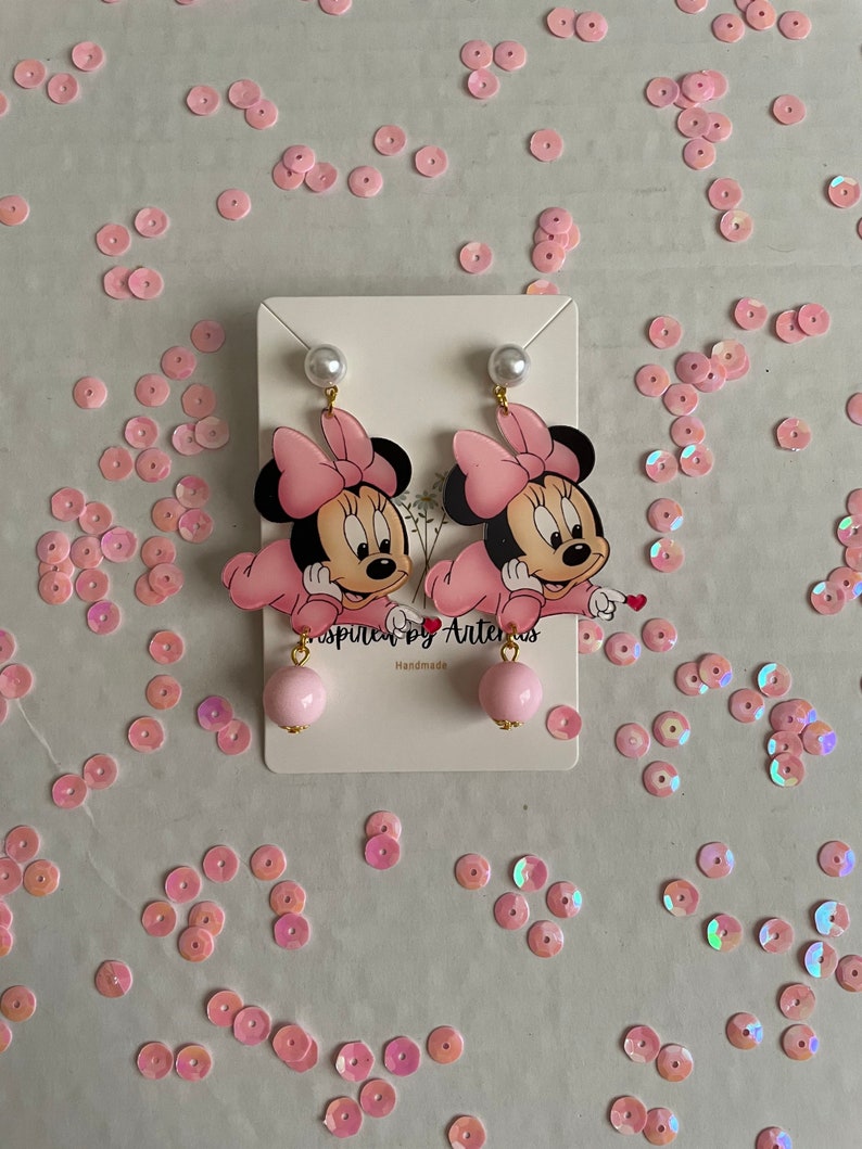 May include: Pink Minnie Mouse earrings with a pink bow and a pearl on top. The earrings have a pink ball at the bottom. The earrings are on a white card with the text "Inspired by Artemis" and "Handmade".