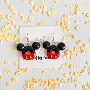 May include: A pair of black and red Mickey Mouse-shaped donut earrings with white accents. The earrings are on a white card with the text "Made by Ari" and "Handmade".