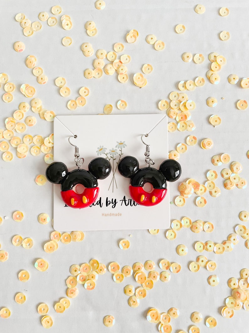 May include: A pair of black and red Mickey Mouse-shaped donut earrings with white accents. The earrings are on a white card with the text "Made by Ari" and "Handmade".