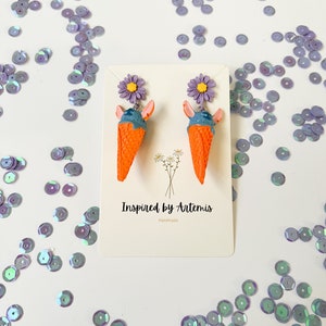 May include: A pair of handmade earrings featuring a blue and orange ice cream cone with a purple flower on top. The earrings are on a white card with the text "Inspired by Artemis Handmade".