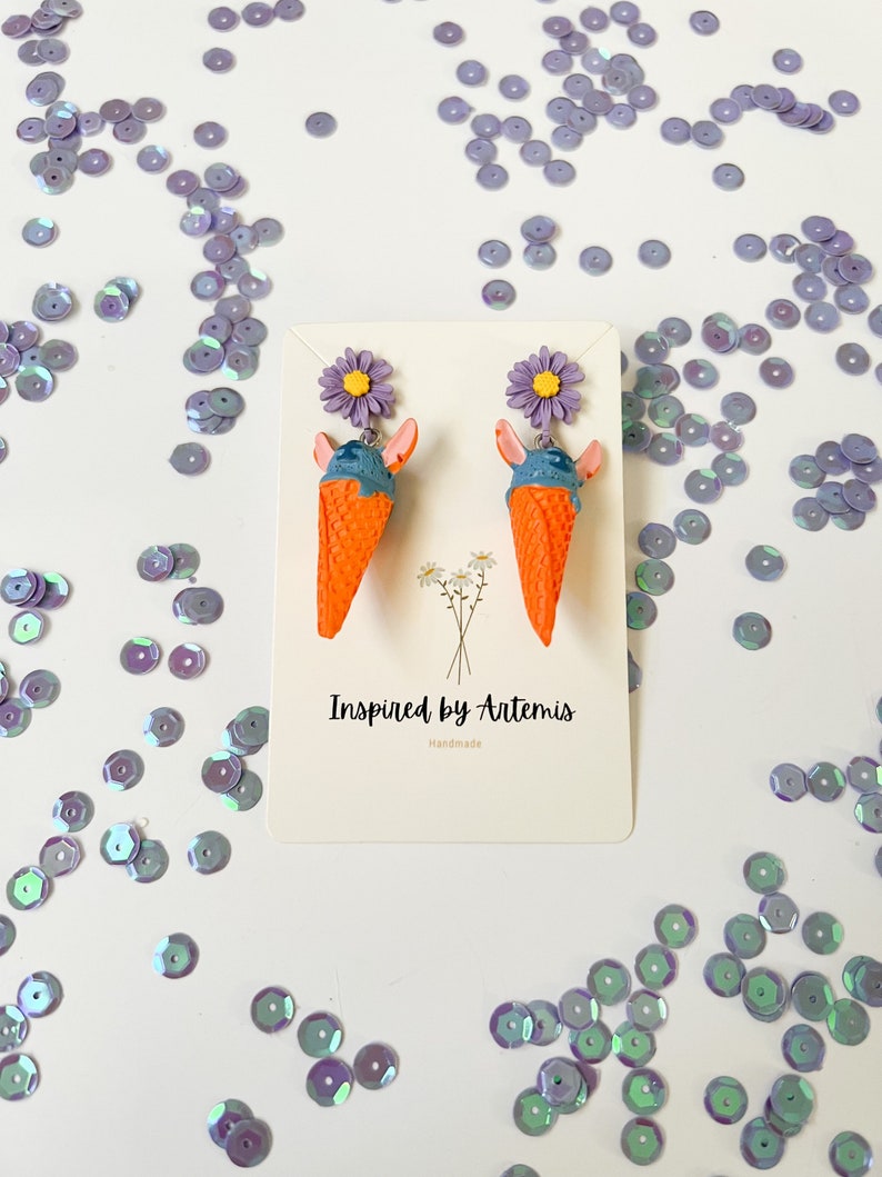May include: A pair of handmade earrings featuring a blue and orange ice cream cone with a purple flower on top. The earrings are on a white card with the text "Inspired by Artemis Handmade".