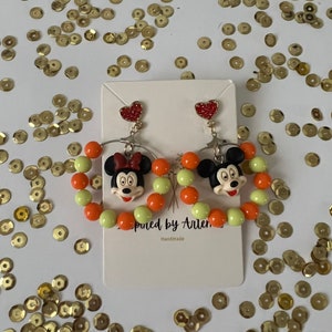 May include: A pair of dangle earrings featuring a cartoon character with a red bow and orange and green beads. The earrings are handmade and have a heart charm at the top.