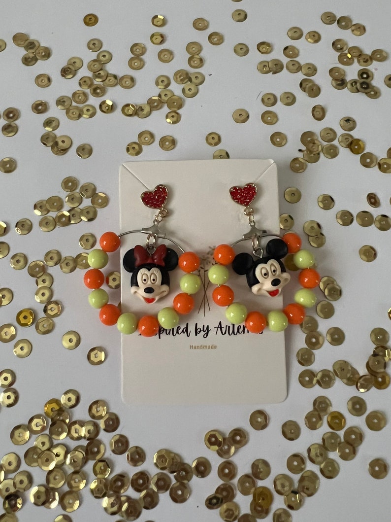 May include: A pair of dangle earrings featuring a cartoon character with a red bow and orange and green beads. The earrings are handmade and have a heart charm at the top.
