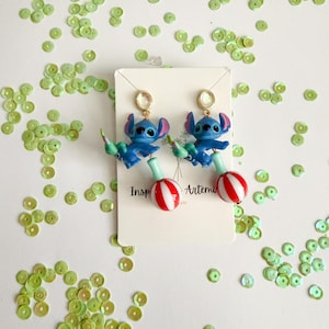 May include: A pair of blue character earrings with red and white striped lollipop-shaped bottoms. The earrings feature a gold-colored stud and are set against a white background scattered with green sequins. The earrings are inspired by a popular animated character.