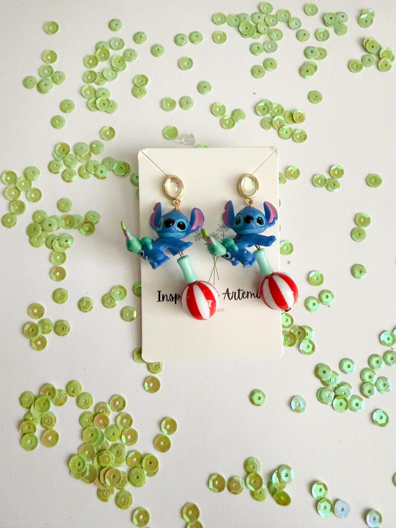 May include: A pair of blue character earrings with red and white striped lollipop-shaped bottoms. The earrings feature a gold-colored stud and are set against a white background scattered with green sequins. The earrings are inspired by a popular animated character.