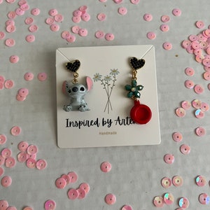 Peut inclure: Une paire de boucles d'oreilles d&eacute;pareill&eacute;es. Une boucle d'oreille pr&eacute;sente un charm en forme de c&oelig;ur noir, une figurine Stitch et une cha&icirc;ne dor&eacute;e. L'autre boucle d'oreille a un charm en forme de c&oelig;ur noir, une fleur verte, un charm rouge et une cha&icirc;ne dor&eacute;e. Les boucles d'oreilles sont sur une carte blanche avec le texte "Inspired by Arte" et "Handmade".