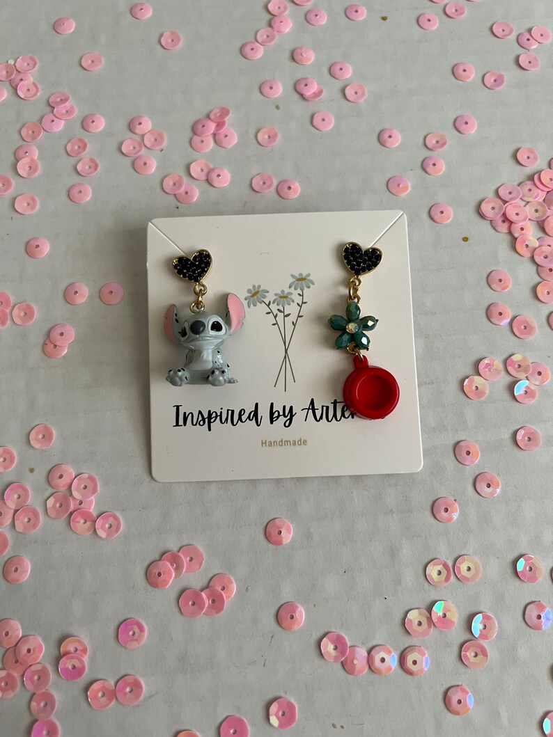 Peut inclure: Une paire de boucles d'oreilles d&eacute;pareill&eacute;es. Une boucle d'oreille pr&eacute;sente un charm en forme de c&oelig;ur noir, une figurine Stitch et une cha&icirc;ne dor&eacute;e. L'autre boucle d'oreille a un charm en forme de c&oelig;ur noir, une fleur verte, un charm rouge et une cha&icirc;ne dor&eacute;e. Les boucles d'oreilles sont sur une carte blanche avec le texte "Inspired by Arte" et "Handmade".