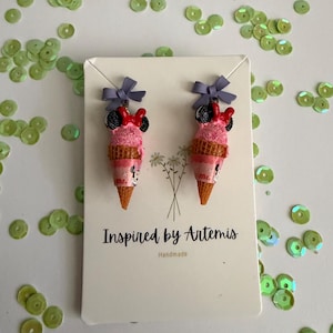 May include: A pair of novelty earrings shaped like pink ice cream cones with Minnie Mouse details. Each cone has a purple bow and a red bow. The earrings are on a white card with the text "Inspired by Artemis" and surrounded by green sequins.