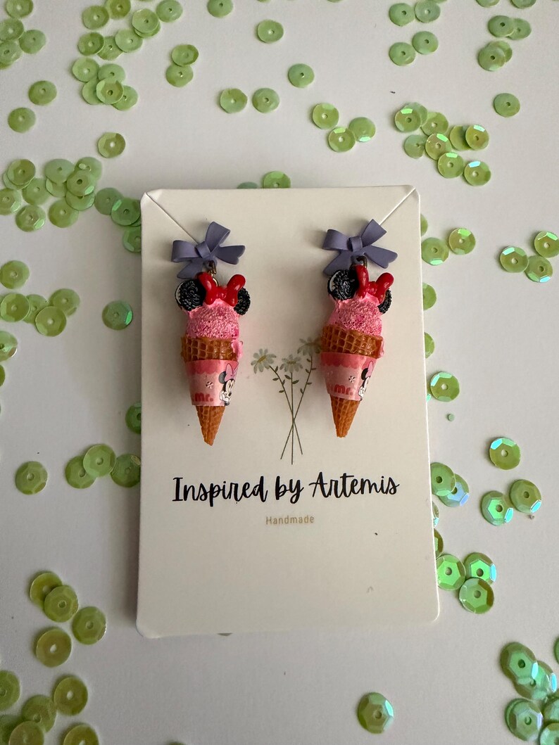 May include: A pair of novelty earrings shaped like pink ice cream cones with Minnie Mouse details. Each cone has a purple bow and a red bow. The earrings are on a white card with the text "Inspired by Artemis" and surrounded by green sequins.