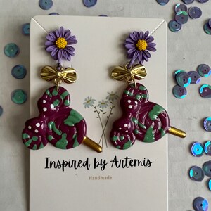 May include: Purple and green swirl lollipop earrings with a gold bow and purple flower charm. The earrings are handmade and the card reads "Inspired by Artemis, Handmade".