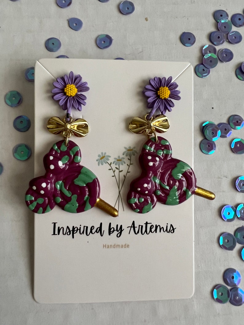 May include: Purple and green swirl lollipop earrings with a gold bow and purple flower charm. The earrings are handmade and the card reads "Inspired by Artemis, Handmade".