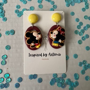 Puede incluir: Un par de pendientes hechos a mano con una imagen de dibujos animados de Mickey Mouse y Minnie Mouse bes&aacute;ndose. Los pendientes est&aacute;n hechos de arcilla polim&eacute;rica y tienen un fondo marr&oacute;n. Los pendientes est&aacute;n en una tarjeta blanca con el texto "Inspired by Artemis" y "Handmade".