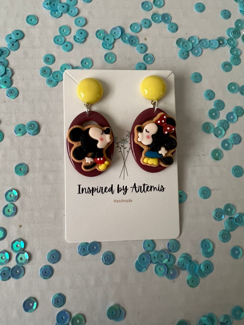 Puede incluir: Un par de pendientes hechos a mano con una imagen de dibujos animados de Mickey Mouse y Minnie Mouse bes&aacute;ndose. Los pendientes est&aacute;n hechos de arcilla polim&eacute;rica y tienen un fondo marr&oacute;n. Los pendientes est&aacute;n en una tarjeta blanca con el texto "Inspired by Artemis" y "Handmade".