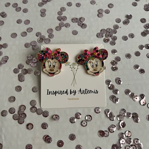 May include: A pair of gold-tone earrings featuring a cartoon character with a pink bow, white face, and colorful rhinestones. The earrings are displayed on a white card with the text "Inspired by Artemis Handmade".