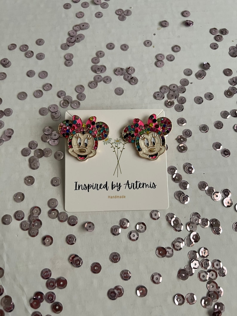 May include: A pair of gold-tone earrings featuring a cartoon character with a pink bow, white face, and colorful rhinestones. The earrings are displayed on a white card with the text "Inspired by Artemis Handmade".