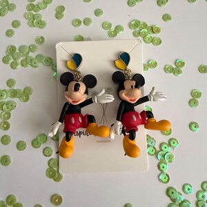 May include: A pair of Mickey Mouse dangle earrings. Each earring features a black Mickey Mouse figure with red shorts, yellow shoes, and white gloves. The earrings have a blue and yellow accent at the top. The card behind the earrings says "Handmade".