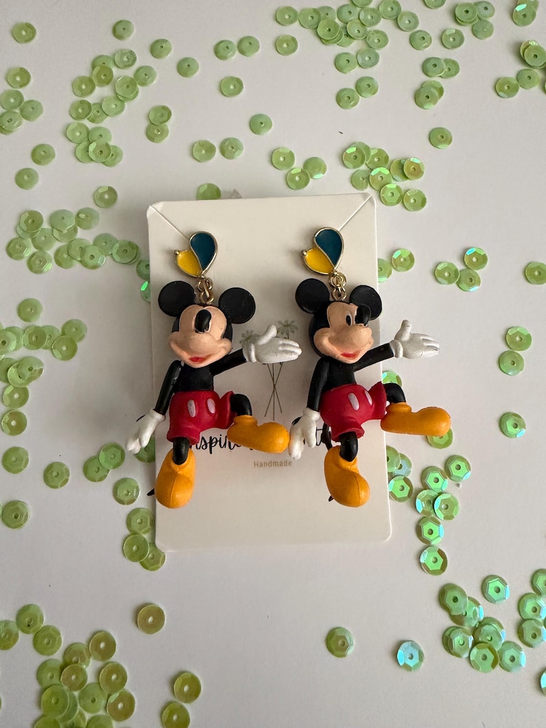 May include: A pair of Mickey Mouse dangle earrings. Each earring features a black Mickey Mouse figure with red shorts, yellow shoes, and white gloves. The earrings have a blue and yellow accent at the top. The card behind the earrings says "Handmade".