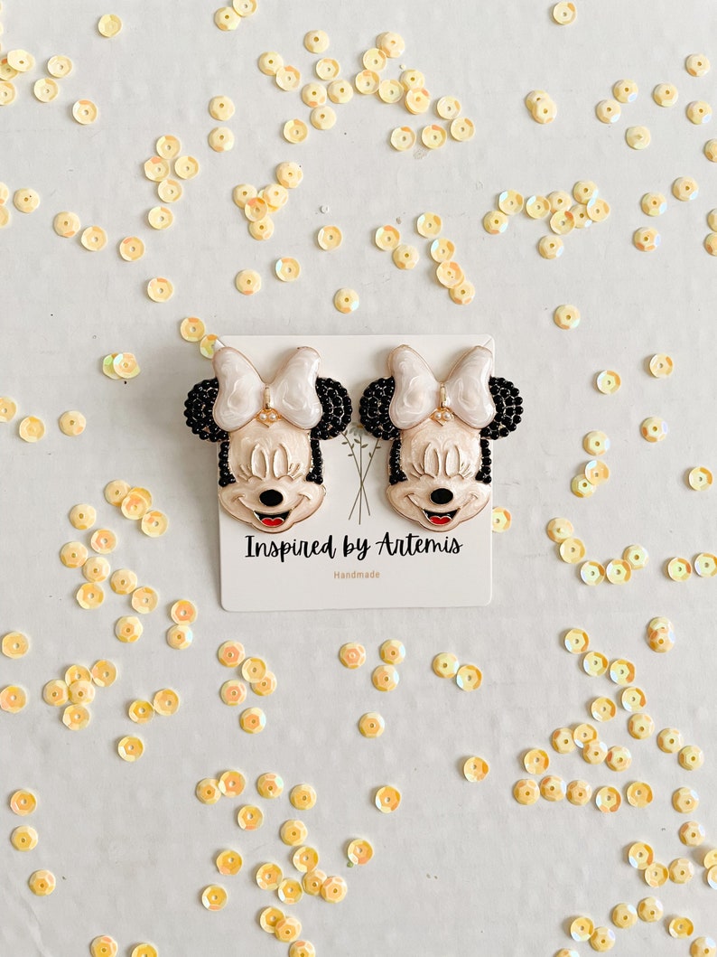 Puede incluir: Un par de pendientes de Minnie Mouse en blanco y negro con detalles de cuentas negras. Los pendientes est&aacute;n hechos a mano y llevan el texto "Inspired by Artemis" y "Handmade".