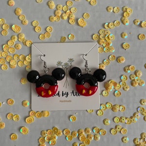 May include: A pair of black and red donut-shaped earrings with Mickey Mouse ears. The earrings are handmade and have a white card behind them with the text "Made by Ari" and "Handmade".