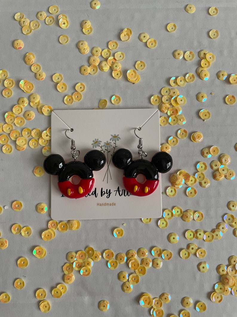 May include: A pair of black and red donut-shaped earrings with Mickey Mouse ears. The earrings are handmade and have a white card behind them with the text "Made by Ari" and "Handmade".