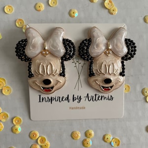 Puede incluir: Un par de pendientes de Minnie Mouse blancos y negros con detalles dorados. Los pendientes est&aacute;n hechos a mano y presentan un lazo blanco, orejas negras y una cara negra con labios rojos. Los pendientes est&aacute;n sobre una tarjeta blanca con el texto "Inspired by Artemis" y "Handmade".