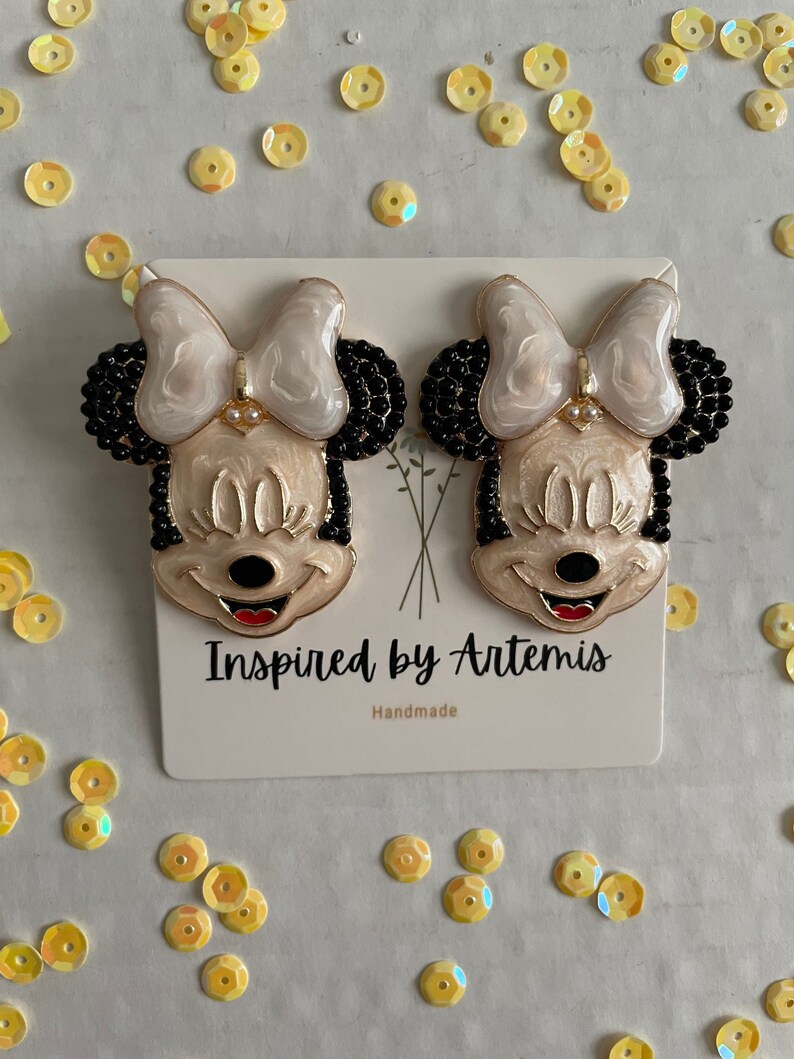 Puede incluir: Un par de pendientes de Minnie Mouse blancos y negros con detalles dorados. Los pendientes est&aacute;n hechos a mano y presentan un lazo blanco, orejas negras y una cara negra con labios rojos. Los pendientes est&aacute;n sobre una tarjeta blanca con el texto "Inspired by Artemis" y "Handmade".