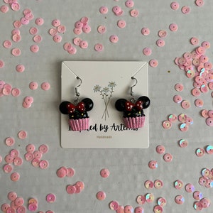 May include: A pair of black and red cupcake earrings with white frosting and a pink bow. The earrings are on a white card with the text "Inspired by Artemis" and "Handmade".