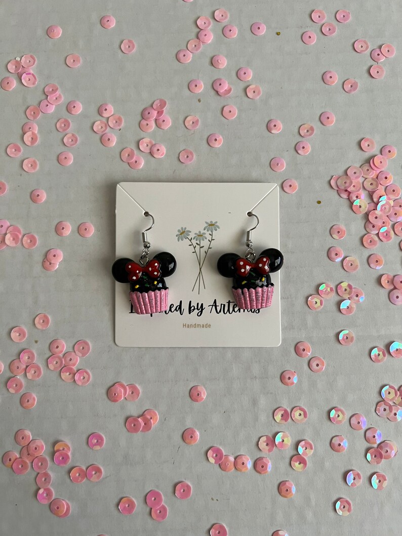 May include: A pair of black and red cupcake earrings with white frosting and a pink bow. The earrings are on a white card with the text "Inspired by Artemis" and "Handmade".