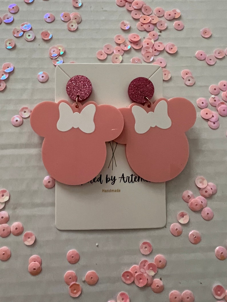 Puede incluir: Pendientes de color rosa en forma de Minnie Mouse con lazos blancos y detalles de purpurina. Hecho a mano.