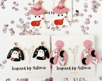 May include: Four pairs of Halloween-themed polymer clay earrings. The first pair features a ghost wearing a pink witch hat. The second pair features a ghost wearing a black and orange sweater. The third pair features a pink ghost with a crescent moon and stars. The fourth pair features a pink ghost with a crescent moon and stars. All earrings are handmade and feature gold accents.