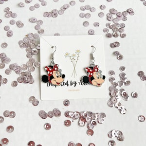 May include: A pair of silver earrings featuring a cartoon image of Minnie Mouse. The earrings are on a white card with the text "Inspired by Ava" and "Handmade".