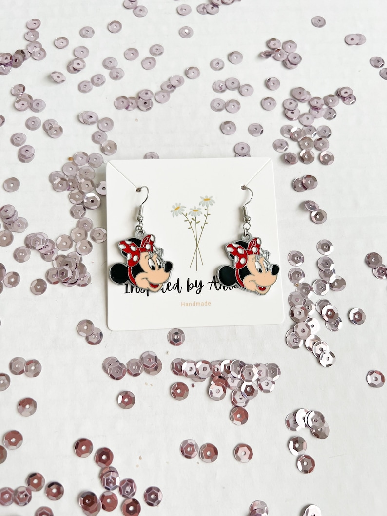 May include: A pair of silver earrings featuring a cartoon image of Minnie Mouse. The earrings are on a white card with the text "Inspired by Ava" and "Handmade".