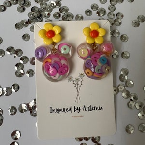May include: A pair of handmade earrings featuring a yellow flower charm and a clear resin charm filled with colorful confetti. The earrings are labeled "Inspired by Artemis" and "Handmade".